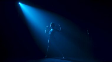 Siluet, esnek, siyah elbiseli genç bir balerin stüdyonun mavi sahne ışıkları altında karanlıkta zarif bir şekilde dans ediyor. Yavaş çekim.