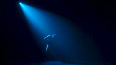 Siluet, esnek, siyah elbiseli genç bir balerin stüdyonun mavi sahne ışıkları altında karanlıkta zarif bir şekilde dans ediyor. Yavaş çekim.