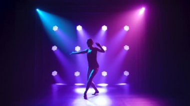 Siyahlar içinde profesyonel bir balerin. Dinamik spot ışıkları altında zarif bir şekilde bale yapıyor ve sahnede sigara içiyor. Güzel, ince bir siluetin silueti. Yavaş çekim.