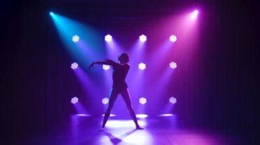 Siyahlar içinde profesyonel bir balerin. Dinamik spot ışıkları altında zarif bir şekilde bale yapıyor ve sahnede sigara içiyor. Güzel, ince bir siluetin silueti. Yavaş çekim.