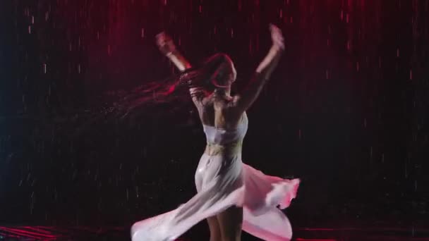 Uma bela jovem em um vestido molhado dança apaixonadamente sob as gotas de chuva. Fundo escuro com luz de estúdio. Movimento lento. Fechar. — Vídeo de Stock