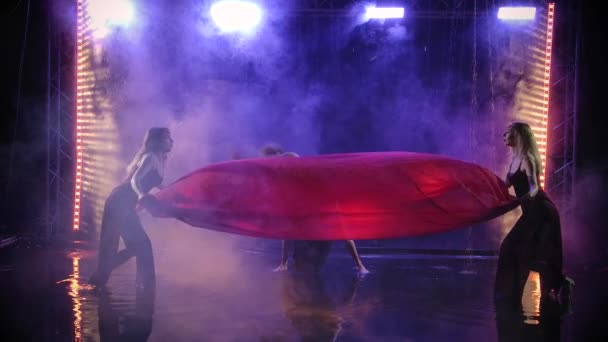 O grupo de dança das mulheres dança graciosamente com um pano vermelho de seda entre as gotas de chuva. Filmado em um estúdio fumegante escuro com iluminação de fundo. Movimento lento. — Vídeo de Stock