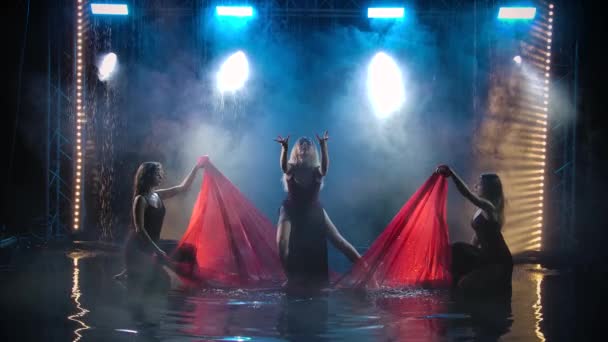Le groupe de danse des femmes danse gracieusement avec un tissu rouge soie parmi les gouttes de pluie. Filmé dans un studio sombre fumé avec rétro-éclairage. Mouvement lent. — Video