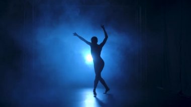 Stüdyonun mavi sahne ışıkları altında karanlıkta esnek dans edebilen siyah elbiseli profesyonel bir balerin. Yavaş çekim.