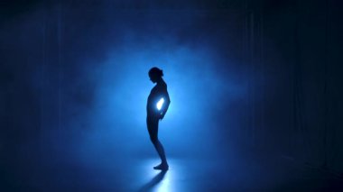 Stüdyonun mavi sahne ışıkları altında karanlıkta esnek dans edebilen siyah elbiseli profesyonel bir balerin. Yavaş çekim.