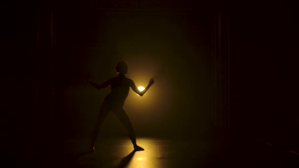 Silhueta uma bailarina flexível fina em bodysuit preto dança graciosa na escuridão abaixo de uns holofotes amarelos teatrais do estúdio. Movimento lento. — Vídeo de Stock