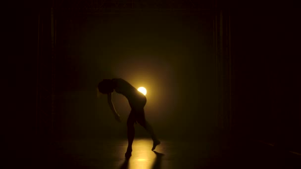 Silhouette μια λεπτή ευέλικτη μπαλαρίνα σε μαύρο κοστούμι σώμα χαριτωμένη χορό στο σκοτάδι κάτω από ένα θεατρικό κίτρινο προβολείς του στούντιο. Αργή κίνηση. — Αρχείο Βίντεο