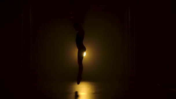 Silhueta uma bailarina flexível fina em bodysuit preto dança graciosa na escuridão abaixo de uns holofotes amarelos teatrais do estúdio. Movimento lento. — Vídeo de Stock