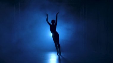 Stüdyonun mavi sahne ışıkları altında karanlıkta esnek dans edebilen siyah elbiseli profesyonel bir balerin. Yavaş çekim.