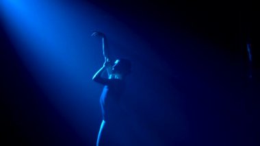 Siluet, siyah takım elbiseli, esnek, genç bir balerin zarif bir dans. Stüdyonun mavi sahne ışıkları altında karanlıkta çekildi. Yavaş çekim.