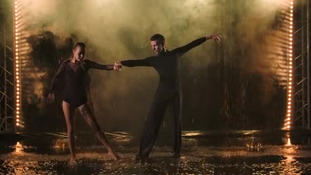 Atractivo hombre y mujer bailan graciosamente jive en los charcos de lluvia bajo las gotas de agua. Fondo retroiluminado ahumado. Movimiento lento. — Vídeo de stock