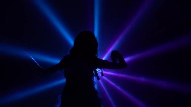 Beyaz doğulu kostümlü, kalçasını sallayan bir dansçı kadın. Duman ve neon ışıkla karanlık bir stüdyoda çekildi. İnce, esnek bir vücudun siluetleri. Kapat..