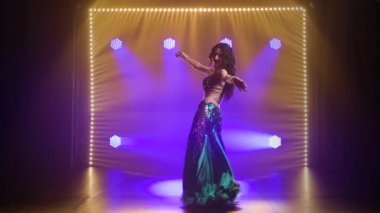 Yeşil, doğulu bir kostüm giyen kız klasik göbek dansı yapıyor. Duman ve mavi neon ışıklarıyla karanlık bir stüdyoda çekildi..