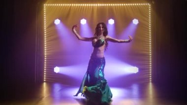 Yeşil, doğulu bir kostüm giyen kız klasik göbek dansı yapıyor. Duman ve mavi neon ışıklarıyla karanlık bir stüdyoda çekildi..