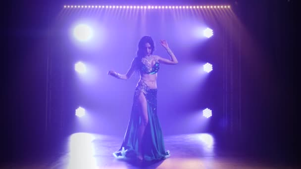 Dívka v zeleném lesklém orientálním kostýmu předvádí klasický břišní tanec. Natočeno v tmavém studiu s kouřem a modrým neonovým osvětlením. — Stock video