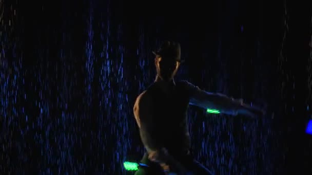 青い光に照らされた水滴を背景に、スタイリッシュな男による光の火のショー。アーティストは夜に光る棒でシルエットになっています。スローモーション。閉じろ!. — ストック動画