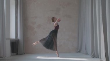 Siyah şeffaf etekli kadın dansçı bale egzersizleri yapıyor. Geniş stüdyodaki pencere kenarındaki dans figürlerinin provaları güneş ışığıyla yıkandı. Yavaş çekim.