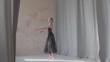 Uzun ışık perdelerinin arkasında siyah şeffaf etekli zarif balerin bir dönüş hareketi sergiliyor. Geniş ve aydınlık stüdyoda dans hareketleri prova ediyor. Yavaş çekim.