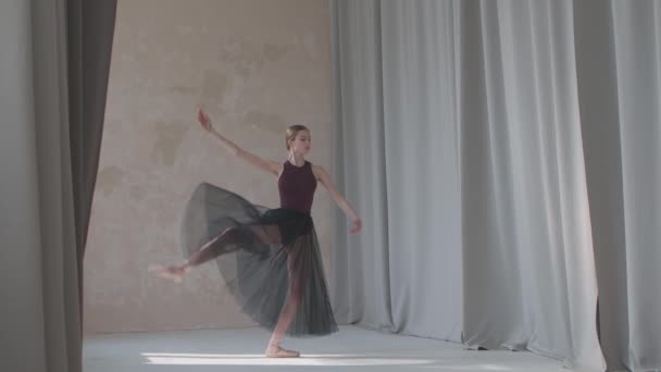 Fragiele jonge ballerina tegen de achtergrond van lange lichte gordijnen tilt haar benen hoog. Repeteert danspasjes in de ruime en helder verlichte studio. Langzame beweging. — Stockvideo