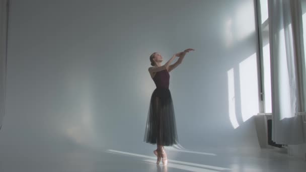 Bailarina en ballet negro tutu vestido bailando pases clásicos en clase cerca de la ventana. Solo calentando antes de la actuación. Fondo blanco de un amplio estudio bañado por la luz del día. Movimiento lento. — Vídeos de Stock