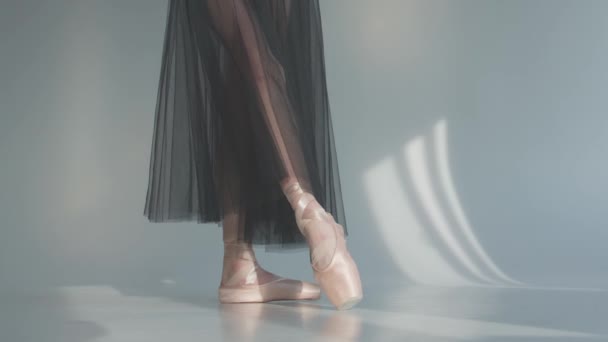 Young ballerina feet as she practices pointe exercises on the white floors. Ballerina in a black transparent skirt shows classic ballet pas. Close up. Slow motion. — Stock Video