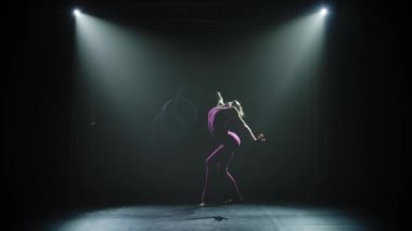 Burgundy mayo giyen zayıf, sevimli, çekici bir sporcu kız kurdeleli ritmik jimnastik elementleri sergiliyor. Karanlık stüdyoda güzel bir ışıkla çekildi. Siluet. Yavaş çekim.