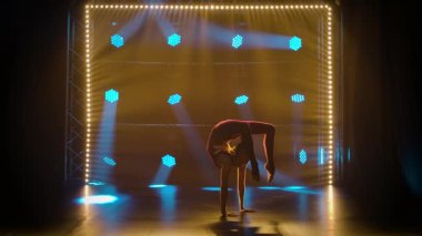 Ritmik bayan jimnastikçi ellerinin üzerinde akrobatik hareketler yapar. Sıska bir jimnastikçi, neon ışıkları olan karanlık bir stüdyoda jimnastik yapıyor. Siluet. Yavaş çekim.