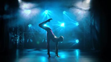 Ritmik bayan jimnastikçi ellerinin üzerinde akrobatik hareketler yapar. Sıska bir jimnastikçi, neon ışıkları olan karanlık bir stüdyoda jimnastik yapıyor. Siluet. Yavaş çekim.