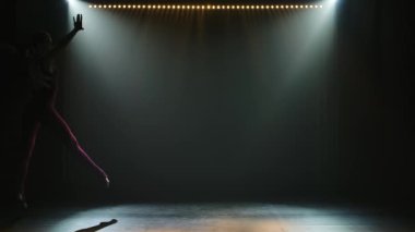 Dar vücutlu genç bayan jimnastikçi akrobatik zıplamalar ve esneme hareketleri yapıyor. Karanlık stüdyoda güzel bir ışıkla çekildi. İnce, esnek bir vücudun silueti. Yavaş çekim.