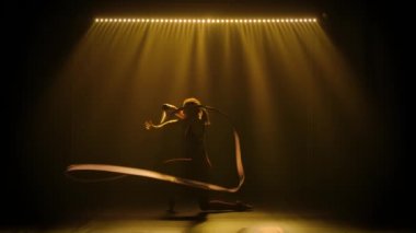 Sporcu kız jimnastikçi, kurdelelerle ve akrobatik egzersizlerle harika bir dans sergiliyor. Sarı neon ışıkları olan karanlık bir stüdyoda çekilmiş. İnce, esnek bir vücudun silueti. Yavaş çekim.