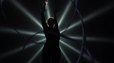 Cılız, genç bir kadın jimnastikçinin silueti sıkı bir vücut içinde jimnastik egzersizini kurdeleyle yapıyor ve esneme hareketleri yapıyor. Alt manzara. Ağır çekim. Kapat..