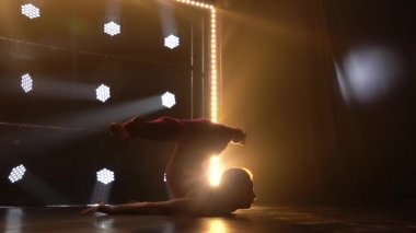 Esnek genç kız jimnastikçi yerde akrobatik bir element sergiliyor. Aktivite konsepti, spor, sağlıklı yaşam tarzı. Siluet. Sarı ışıklı karanlık bir stüdyo. Ağır çekim. Kapat..