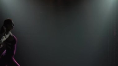 Dar vücutlu ritmik jimnastikçi akrobatik atlayışlar yapar. Sahnelenmiş ışıkla karanlık stüdyoda çekildi. İnce, esnek bir vücudun silueti. Ağır çekim. Kapat..