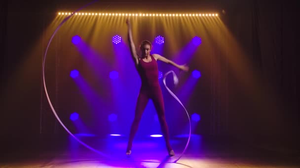 Gymnaste féminin rythmique dansant avec ruban coloré. Gymnaste mince dans le corps bordeaux effectue l'exercice de gymnastique avec un ruban dans un studio sombre avec des néons. Silhouette. Mouvement lent. — Video