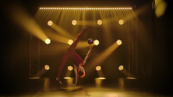 Ginasta feminina rítmica realiza movimentos acrobáticos com uma bola. Ginasta magro em corpo bordô executa o exercício de ginástica em um estúdio escuro com luzes de néon. Silhueta. Movimento lento. — Vídeo de Stock