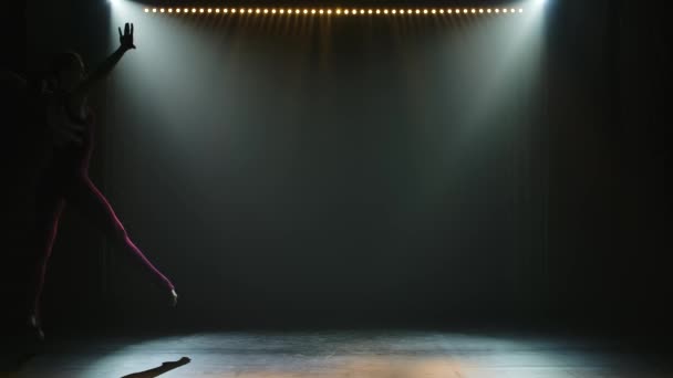 Štíhlá mladá gymnastka v těsně padnoucím těle provádí akrobatické skoky a protahování. Natočeno v tmavém ateliéru s krásným inscenovaným světlem. Silueta štíhlého pružného těla. Zpomalený pohyb. — Stock video