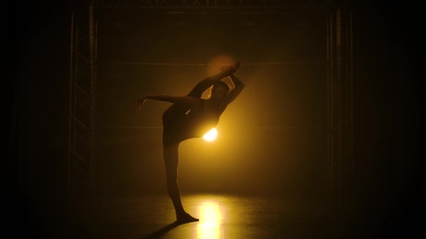 Een turnster voert een acrobatisch element uit op de vloer. Het concept van ritme, sport, gezonde levensstijl. Donkere studio met prachtig geënsceneerd licht. Silhouet slanke flexibele lichaam. Langzame beweging. — Stockvideo