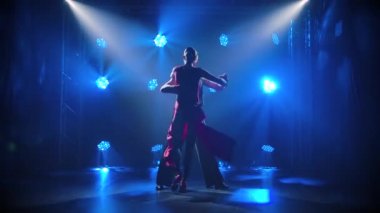 Bir çift tango sanatçısı, koyu mavi arka ışıklandırma stüdyosunda tutkuyla el ele tutuşarak dans ediyor. Ağır çekimde dans eden dansçıların siluetleri.
