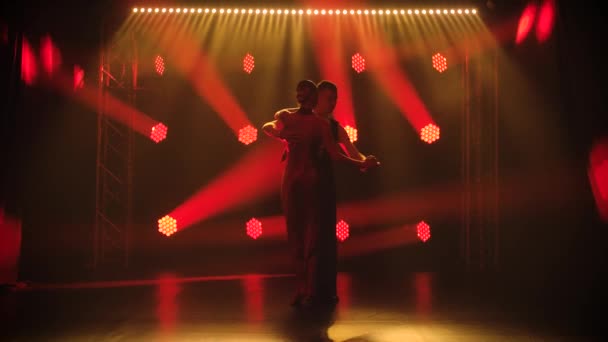 Lady in Lacy avondjurk dansen met man hete Argentijnse tango demonstreren elegante wendingen in donkere studio. Een silhouet van koppels tegen een achtergrond van rode neon lichten. Langzame beweging. — Stockvideo