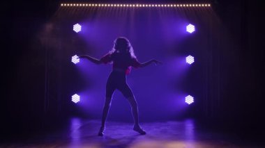 Genç sportif kız aktif olarak funky jazz dansı yapıyor. Karanlık bir stüdyoda arka plandaki mavi ışıklara karşı bir siluet. Yavaş çekim.