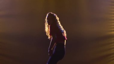 Kıvırcık saçlı bir kız stüdyoda ışıkların arkasında neşeli bir şekilde caz dansı yapıyor. Siluet yakın çekim.