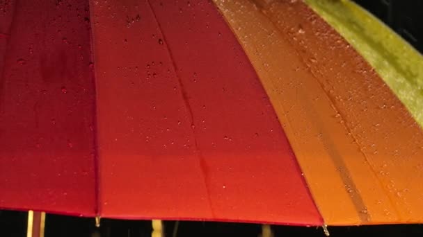 Jasna tęczowa parasolka w deszczu w zamkniętym ciemnym studio. Nagrane z bliska w zwolnionym tempie. — Wideo stockowe