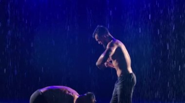 Denge pozisyonunda iki kaslı erkek akrobat. Stüdyoda yağmur damlaları ve su damlaları arasında karmaşık bir akrobatik gösteri sergilendi. Ağır çekim. Kapat..