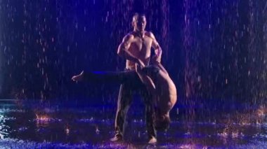 Denge pozisyonunda iki kaslı erkek akrobat. Stüdyoda yağmur damlaları ve su damlaları arasında karmaşık bir akrobatik gösteri sergilendi. Ağır çekim. Kapat..