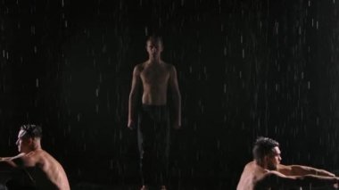 Üç akrobattan oluşan bir grup yağmurda dengelemek için zor numaralar yapıyor. Erkekler arka planda ağır çekimde siyah ışığa karşı dururlar. Kapat..