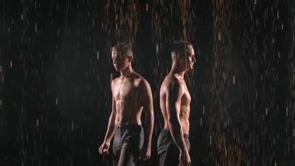 Dos acróbatas gimnásticos en equilibrio posan bajo corrientes de lluvia. Hombres atléticos fuertes realizando trucos difíciles en el trabajo en equipo. Filmado sobre fondo negro con iluminación en cámara lenta. De cerca.. — Vídeos de Stock