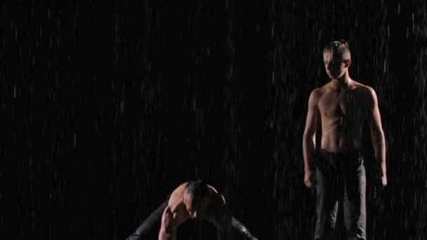 Dua akrobat jantan melakukan handstands di tengah hujan. Kerja sama tim terkoordinasi dengan baik. Difilmkan pada latar belakang hitam dengan iluminasi dalam gerakan lambat. Tutup.. — Stok Video