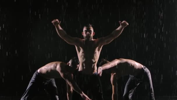 Un grupo de tres acróbatas realizan difíciles trucos de equilibrio bajo la lluvia. Los hombres se paran contra el fondo negro retroiluminado en cámara lenta. De cerca.. — Vídeo de stock