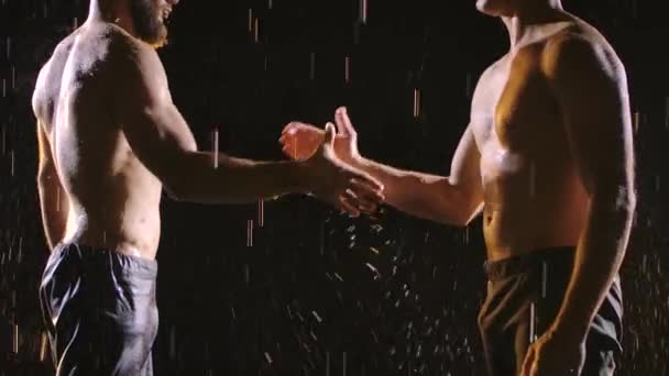Två muskulösa män skakar hand i det stänkande regnet. Bild tagen i studion på svart bakgrund. Sakta i backarna. Närbild. — Stockvideo