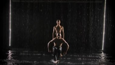 İki genç sporcu amuda kalkıyor. Erkek akrobatlar. Siyah arka planda, su akıntılarının altında çıplak bir gövde. Yavaş çekim.